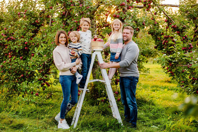 Mom and dad with their two sons and daughter standing on a white ladder in an apple orchard for their fall family photography session in Burlington, ON