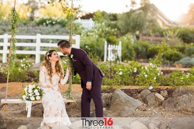 Groom leans in for a kiss as his Bride sits on a swinging bench at the Forever and Always Farm wedding venue in Murrieta, CA