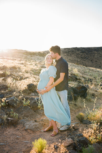 expecting mom and dad snuggle close together on mountain with volcanic rock in st. george utah for maternity photos