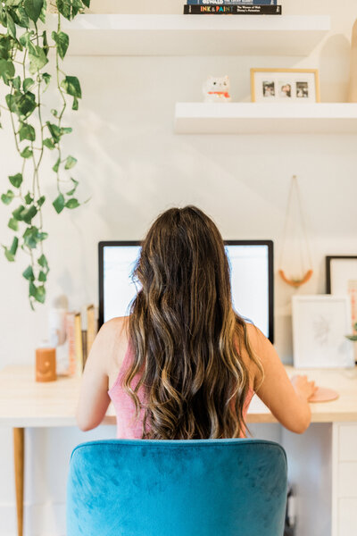 woman with long, dark highlighted hair sitting in a blue velvet desk chair working at an iMac; bohemian office design; hanging plant; floating shelf vignette