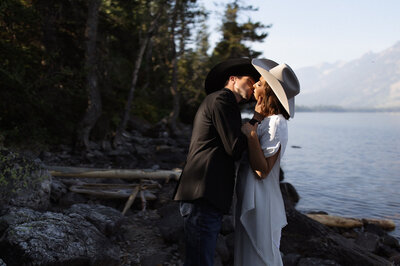 Bride and groom kissing in the forest, groom wearing cowboy hat, captured during a rustic domestic elopement.
