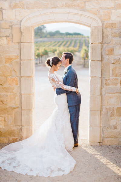 Old Mission Santa Ines Ceremony with Glamorous Sunstone Villa Reception - portrait of a bride and groom embracing under a stone archway that looks like it could be somewhere in Tuscany.