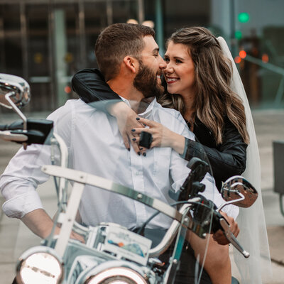 bride and groom on the back of a motorcycle look at each other and smile while bride wraps her hands around his chest