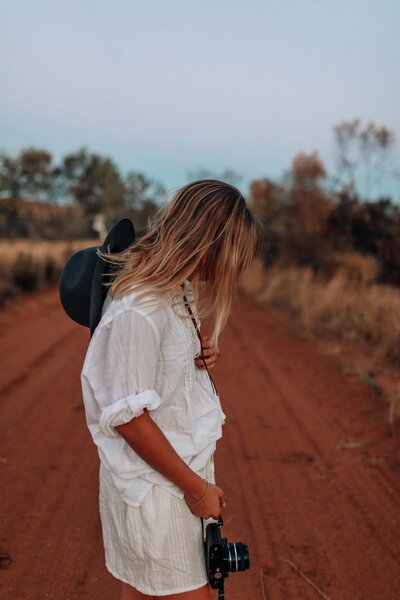 A white woman wearing all white looks down so you cannot see her face on a dirt road holds a camera and a boho hat