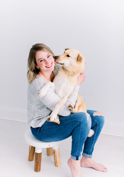 Raleigh photographer Valerie Worth poses in white studio with dog licking her cheek
