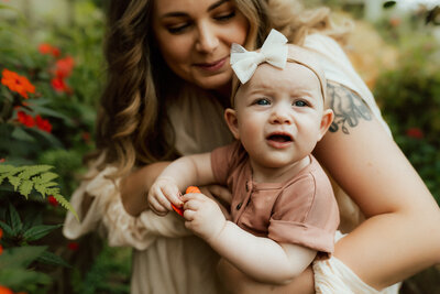 A woman with long hair and a tattoo on her arm holds a baby dressed in a pink outfit and headband with a bow, while surrounded by greenery and red flowers.