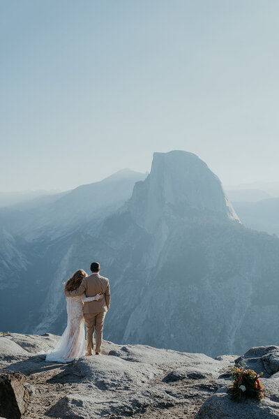 Bride and groom facing away from the camera, looking at Half Dome in the background. Elopement photograph taken at Glacier Point in Yosemite National Park.