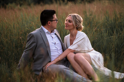Engaged couple look at each other with love, sitting in tall grass near Edmonton, Alberta
