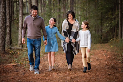 A family of four is holding hands and walking through a forest while they look at each other and giggle.