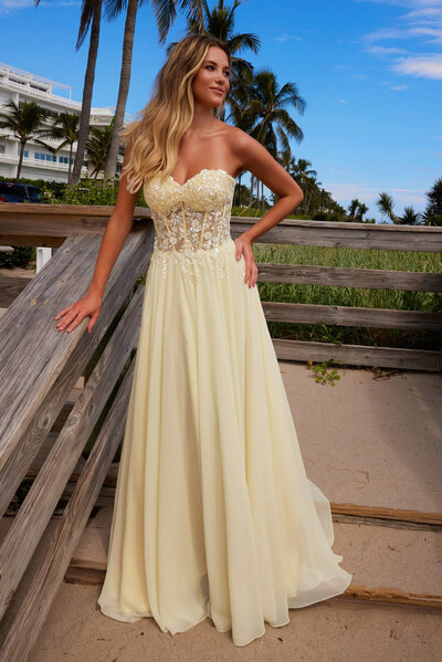 Woman wearing a strapless yellow gown with a sheer embellished bodice near a beach walkway