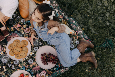 Young girl in denim overalls sitting cross-legged on a picnic blanket outdoors, surrounded by fruit bowls, cookies, and bread.