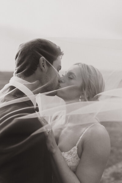 A black and white image of a couple kissing during their sunset portraits, the bride's veil gently flowing around them, creating an intimate and romantic moment that captures the beauty and connection between them.