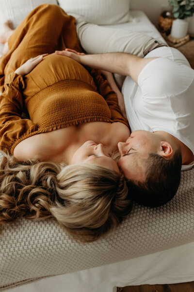 A maternity photo of a pregnant mother lying on a bed next her to husband who is rubbing her belly in a Vancouver Washington natural light studio