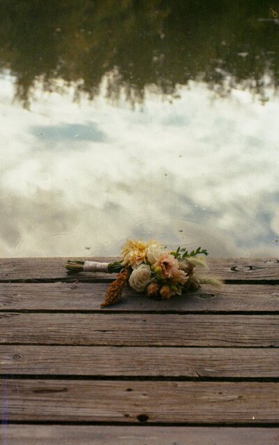 A 35mm film image of a bridal bouquet sitting on the edge of a dock with reflections in the water.