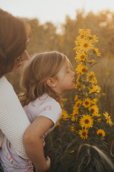 Toddler smelling yellow flowers, while mom is holding her
