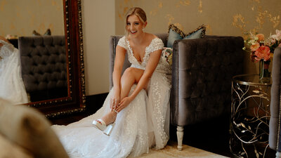 Bride getting ready in wedding suite, sittng in a chair and strapping her heels
