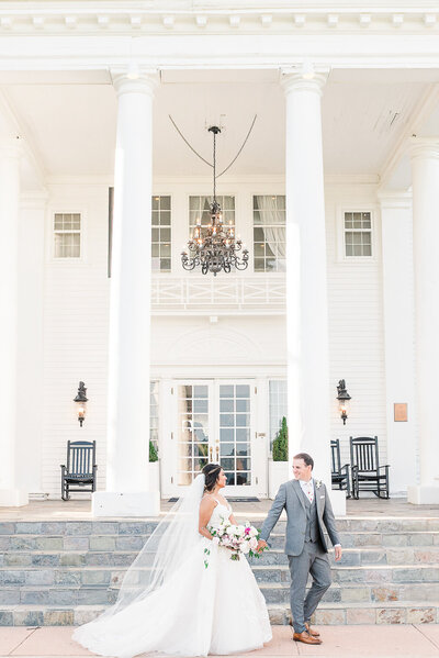 Groom leading bride by the hand in front of a majestic white manor house on their wedding day.