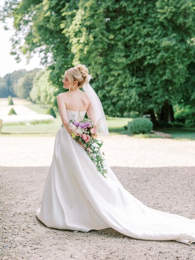 bride standing in her wedding gown her back to the camera showing off her bouquet to aurore bridges Table of Honnor in the gardens of the Château de champlâtreux surrounded by trees with brown chairs and pink linen as wells as yellow and pink floral arrangements