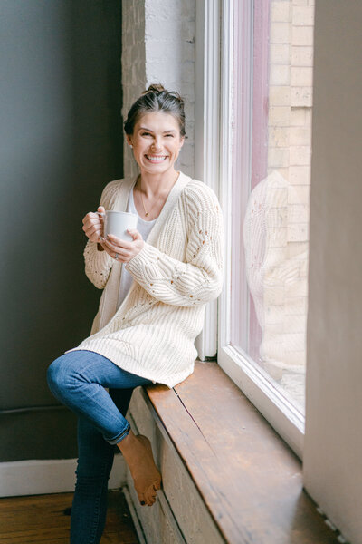 Woman sits on windowsill holding coffee mug while smiling