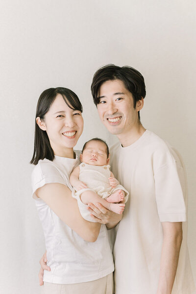 ホワイトコーデでおしゃれな、ママとパパが笑っているニューボーンフォト。赤ちゃんはニューボーンのニットの衣装を着て寝ている。