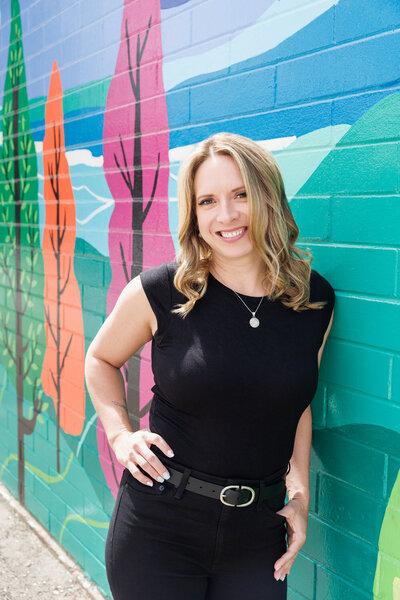 A woman in a black outfit smiles confidently while leaning against a colourful mural in Calgary’s Beltline district, part of the BUMP project. A vibrant and modern take on professional headshots.