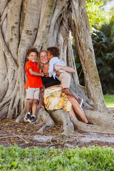 Two young boy's kiss their mom's cheek while she sits on the roots of a banyon tree