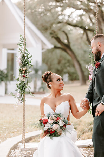 Photo of Texas bride and groom gazing into each other's eyes as bride sits on floral swing