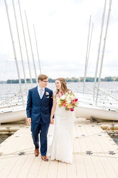 Couple hold hands while walking down pier with sailboats in  background for White Bear Lake Wedding.