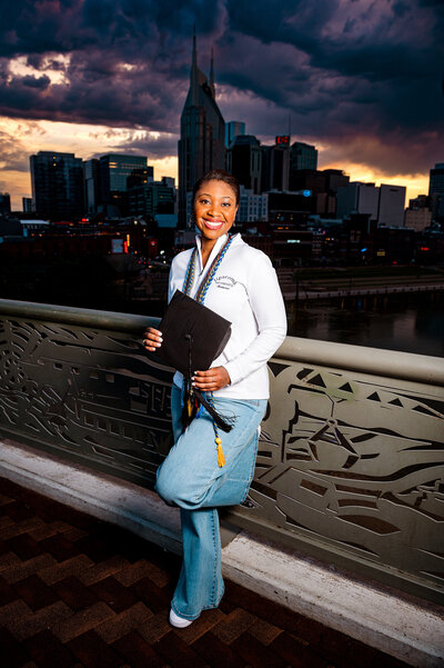 Lipscomb University graduating accounting senior wearing cords and holding cap standing on pedestrian bridge at sunset