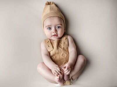 A woman with long, dark curly hair, wearing an orange shirt tied at the waist and dark jeans, smiles at the camera. Pampas grass in a vase is to her left