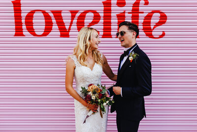 Stylish bride and groom laughing in Digbeth against a pink shutter, with a 'LoveLife'  sign in the background.