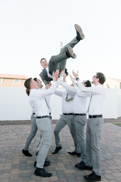 Groomsmen wearing white button down shirts and gray pants throwing groom in the air while laughing.