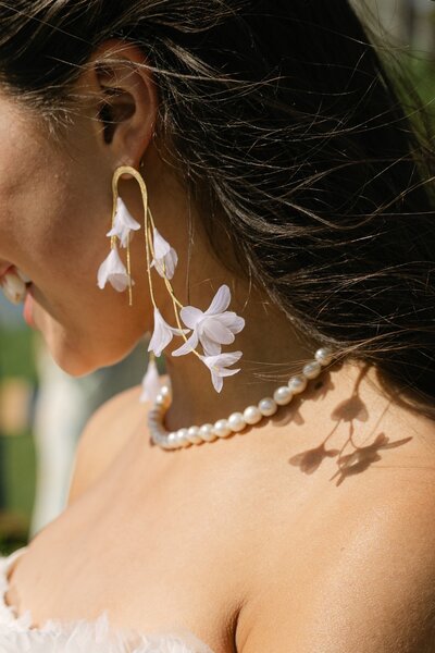 Close-up bridal detail of white floral earrings and pearl necklace captured in natural sunlight by Colorado wedding photographer