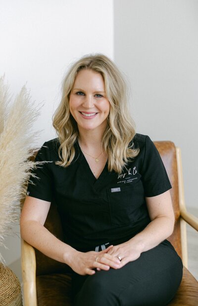 A woman with blonde hair wearing black scrubs sits smiling on a brown chair in a bright room at Falmouth Maine Medical Aesthetics, with pampas grass visible to the side.