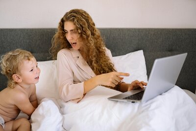 A mom in bed on her laptop with her toddler next to her. 