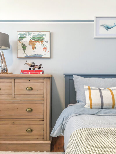 Cozy bedroom designed by a Newcastle interior designer, featuring a wooden dresser, world map, books, toy plane, and bed with blue and gray linens.