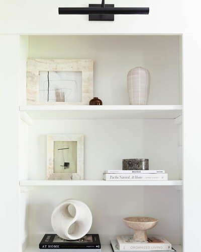 Styled built-in shelf by Deni + Dove Interiors, a luxury NJ interior designer serving Summit, Maplewood, and Montclair. Features curated neutral decor, minimalist artwork, sculptural objects, and design books—showcasing elevated, modern interior styling with a refined, artistic touch.