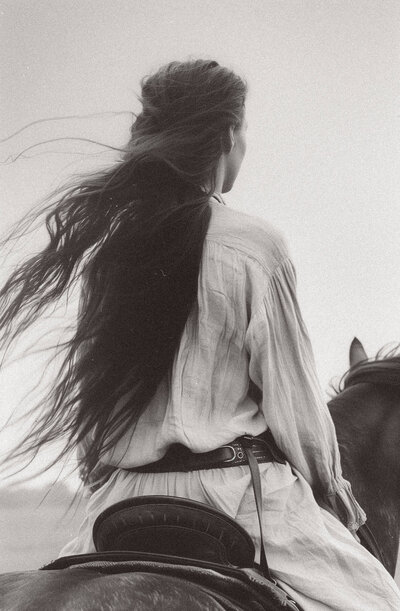   A person with long, flowing hair sits on a saddled horse, facing away from the camera, wearing a loose, light-colored shirt—capturing the serene essence often featured in Sweet Pea Ink Creative’s brand design.