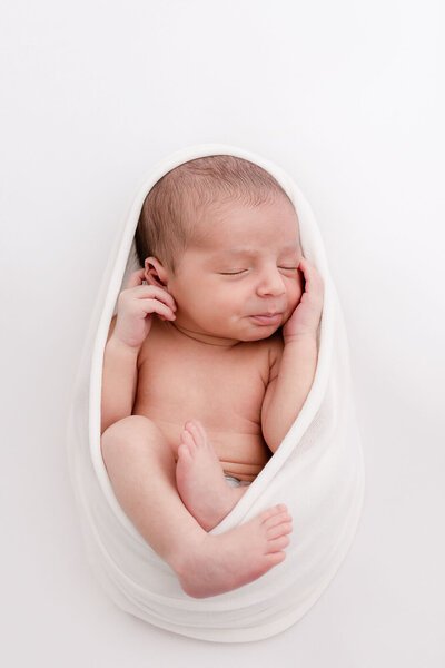 Newborn baby wrapped in a white swaddle with legs hanging out. Hands are up by his face and he is sleeping peacefully on a white backdrop.
