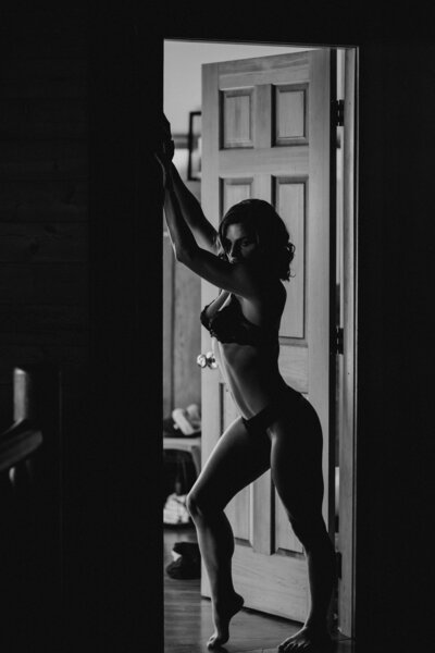 A black and white photo of an athletically built figure stands in a doorway. The light comes from inside the room, leaving the exterior door frame and her silhouette a dark shadow.