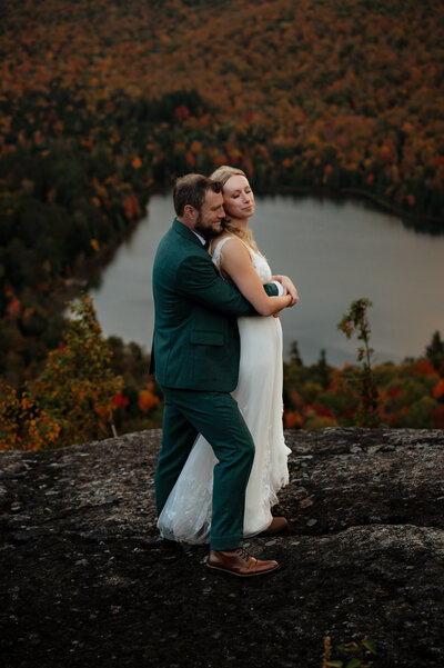 Couple embraces each other on a  ledge in the adirondack mountains with heart lake in the background and fall foliage. 