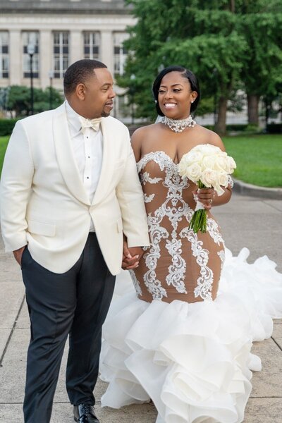 Bride and groom walking hand in hand in downtown Indianapolis after wedding at Indianapolis War Memorial, featuring bride in a couture lace gown by Vivien Jackson with dramatic ruffles and groom in classic white tuxedo.