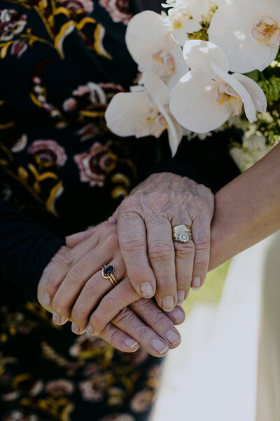 The grandmothers hands grasp the brides hand and both their wedding rings are on display. You can also see the brides spring bouquet 