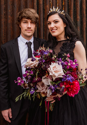 Gothic bride and groom, bouquet full deep purple and red flowers by the little flower hut