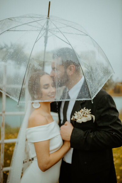 groom kisses bride as they huddle under their clear umbrella shielding from the rain