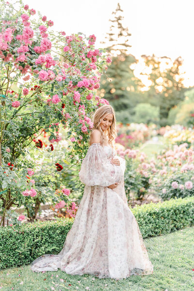 An expecting mother stands in a rose garden dressed in a long pink floral gown holding her growing baby bump photographed by bay area photographer, Light Livin Photography.
