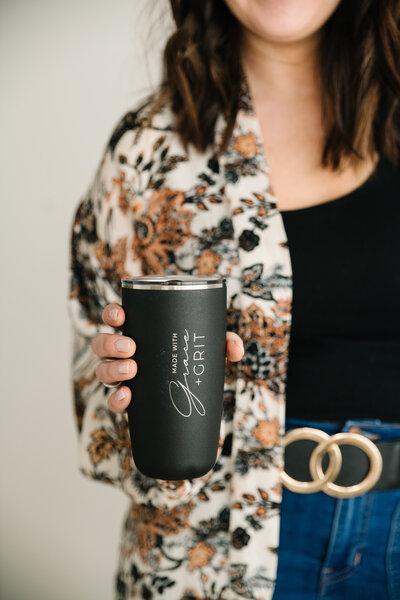 A woman holds a coffee tumbler with the Made with Grace and Grit logo