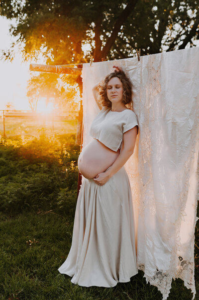 A bare belly pregnant woman in a two piece ivory set standing next to a clothes line at little glass shack in Howe, texas