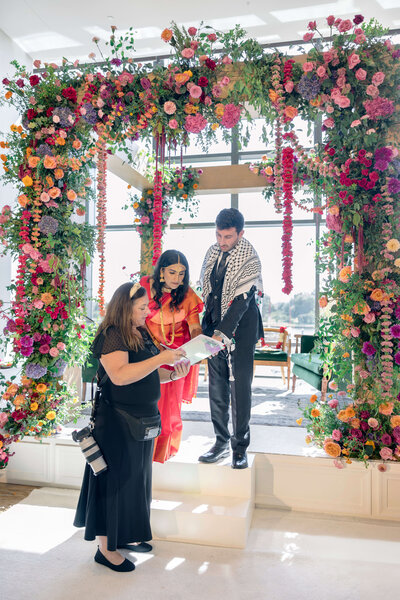 A couple stands on a small platform under a floral arch, dressed in formal attire. A woman with a camera shows them something on a digital screen. Bright, colorful flowers adorn the arch and the room is filled with natural light.