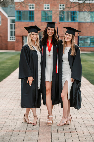 Three Quinnipiac University graduates in black caps and gowns pose together on campus, celebrating their achievement with smiles and confidence. Captured during a Connecticut college graduation photography session by Nicole Mele Photography, highlighting friendship and success.